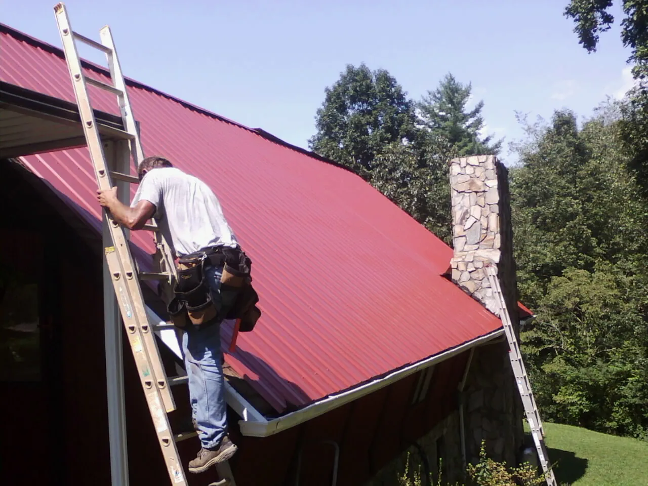 Roofer on a ladder beside a red metal roof and stone chimney — Soffit & Fascia Repair by Roofing Brothers in Flemington